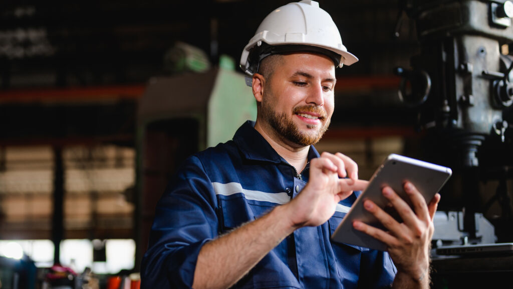 Homem com uniforme industrial e capacete utilizando um tablet em um ambiente de fábrica, sorrindo enquanto interage com o dispositivo.