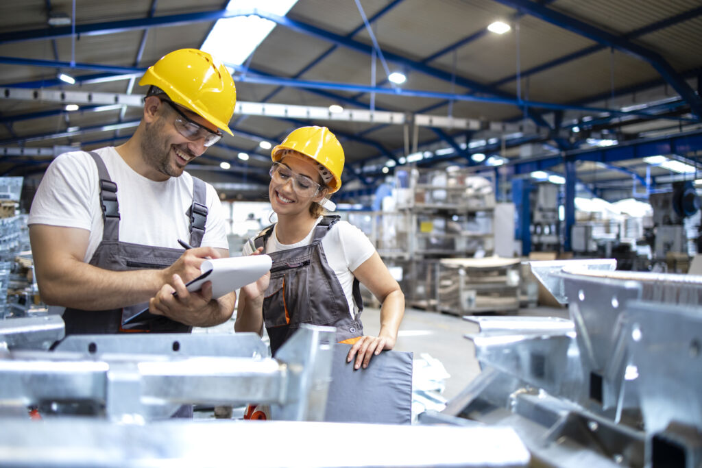 Dois trabalhadores sorridentes em um ambiente industrial, usando capacetes de segurança e óculos de proteção, analisando documentos em uma prancheta enquanto estão cercados por peças metálicas em um grande galpão.