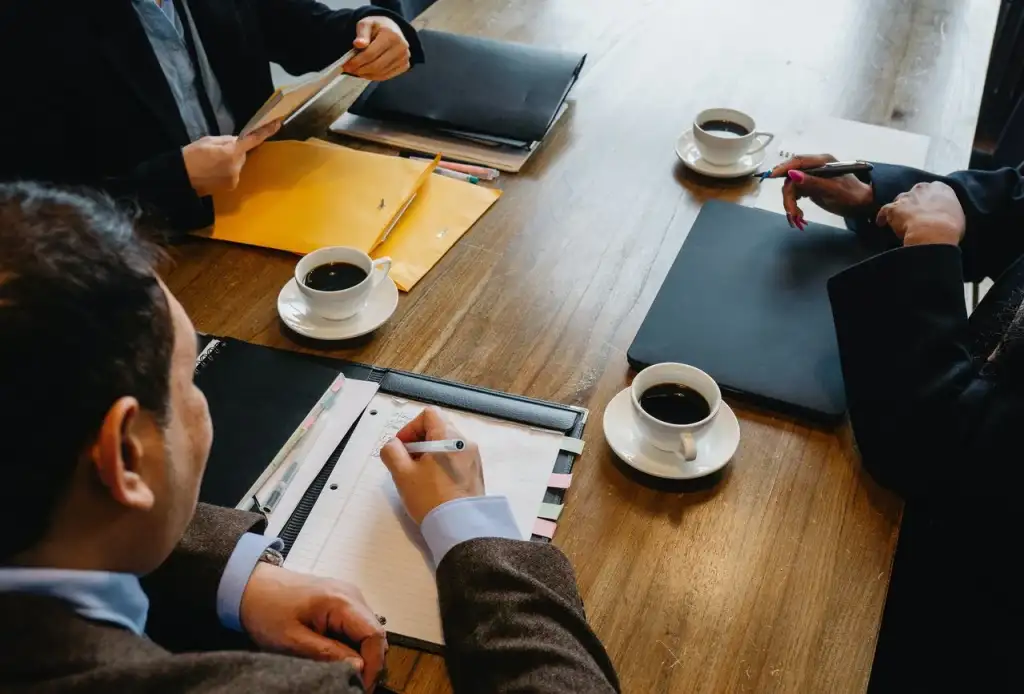 mesa de reuniões com três pessoas ao redor e três xícaras de café e fichário em cima