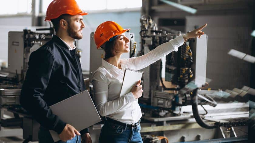 Dois profissionais usando capacetes laranja e roupas de trabalho em uma planta industrial. A mulher aponta para uma área específica da fábrica enquanto segura um bloco de notas, e o homem ao lado dela segura um laptop, observando atentamente. Ao fundo, há máquinas e equipamentos industriais.