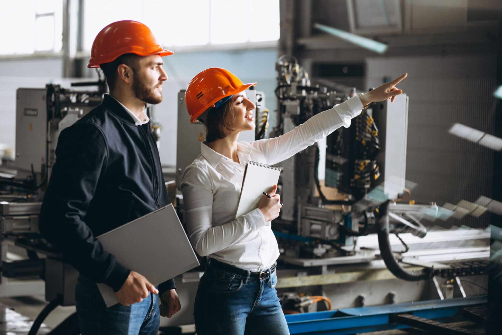 Dois profissionais usando capacetes laranja e roupas de trabalho em uma planta industrial. A mulher aponta para uma área específica da fábrica enquanto segura um bloco de notas, e o homem ao lado dela segura um laptop, observando atentamente. Ao fundo, há máquinas e equipamentos industriais.