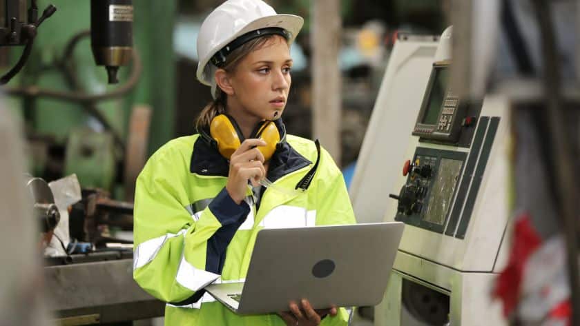 Engenheira usando capacete de segurança e equipamento de alta visibilidade enquanto opera uma máquina industrial. Ela segura um laptop e usa protetores auriculares, indicando monitoramento técnico ou controle de processos em uma fábrica.
