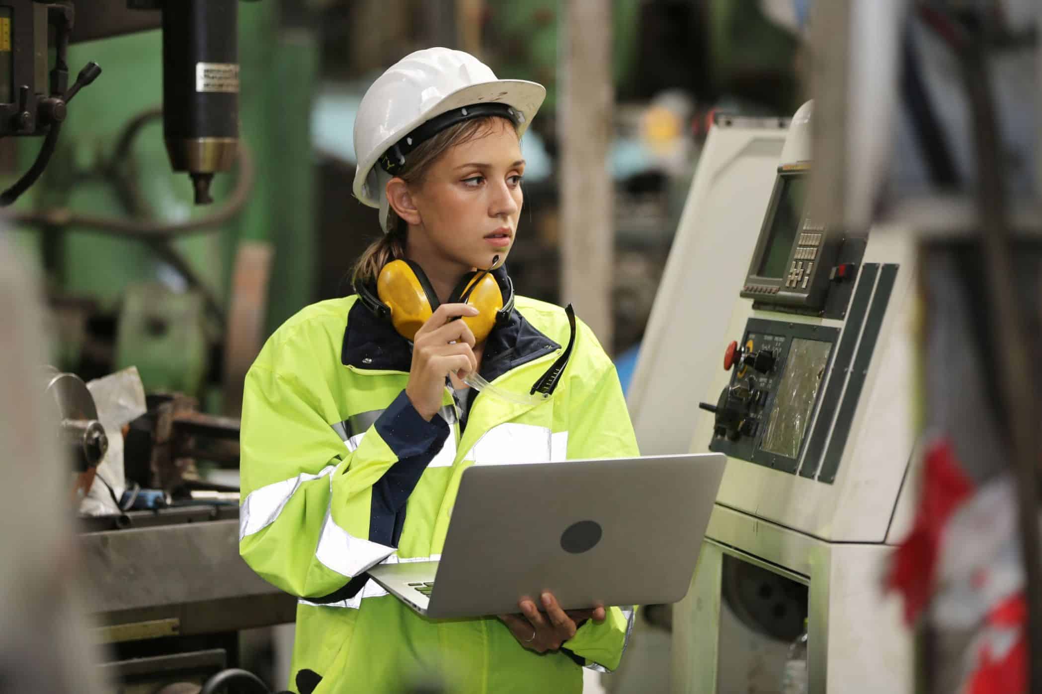 Engenheira usando capacete de segurança e equipamento de alta visibilidade enquanto opera uma máquina industrial. Ela segura um laptop e usa protetores auriculares, indicando monitoramento técnico ou controle de processos em uma fábrica.