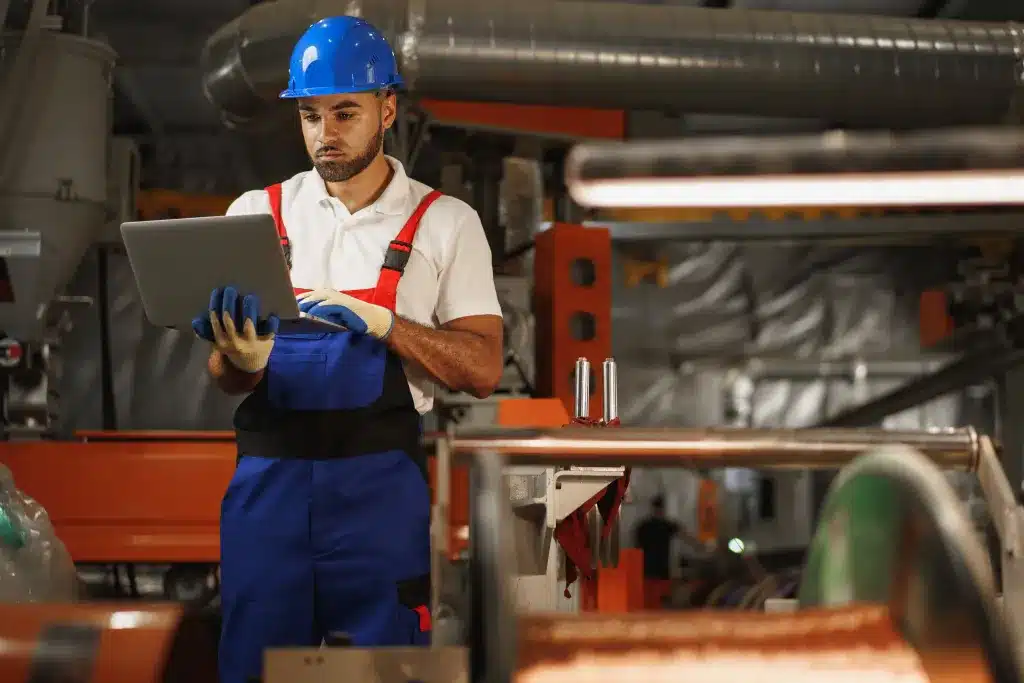 planejamento-e-controle-da-manutencao-homem-jovem-no-chao-de-fabrica-com-capacete-segurando-na-mao-um-notebook