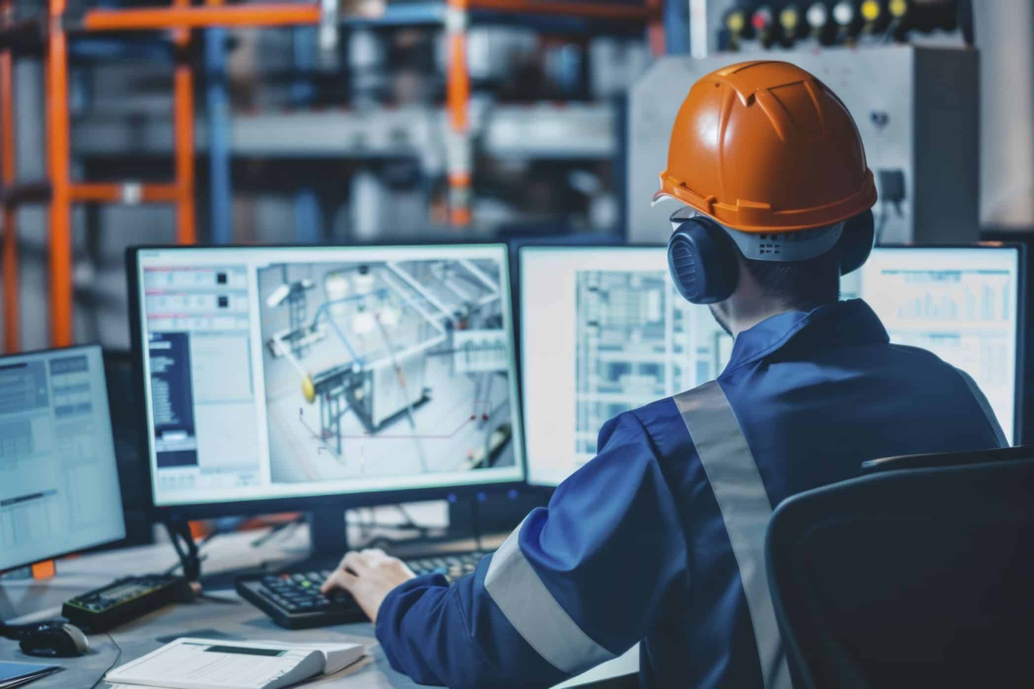 Profissional industrial usando capacete de segurança e protetores auriculares, monitorando painéis de controle e gráficos em várias telas de computador, em um ambiente de fábrica.