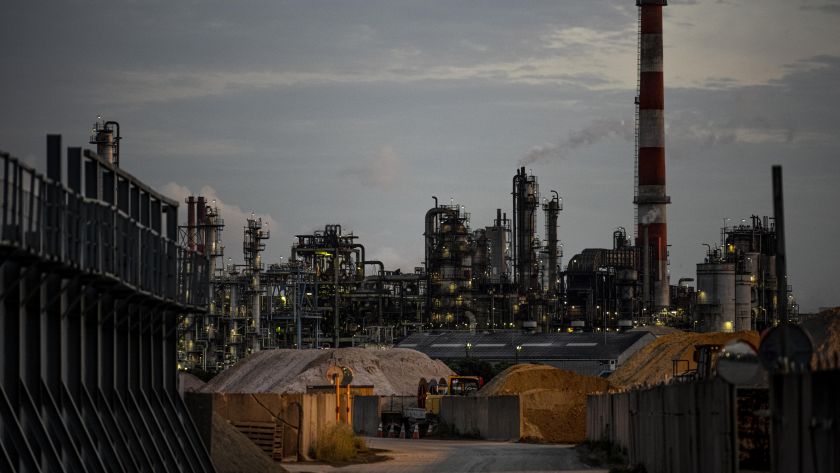 Vista de uma fábrica industrial ao entardecer, com diversas torres e estruturas de produção iluminadas, pilhas de materiais no primeiro plano e uma chaminé ao fundo emitindo fumaça.