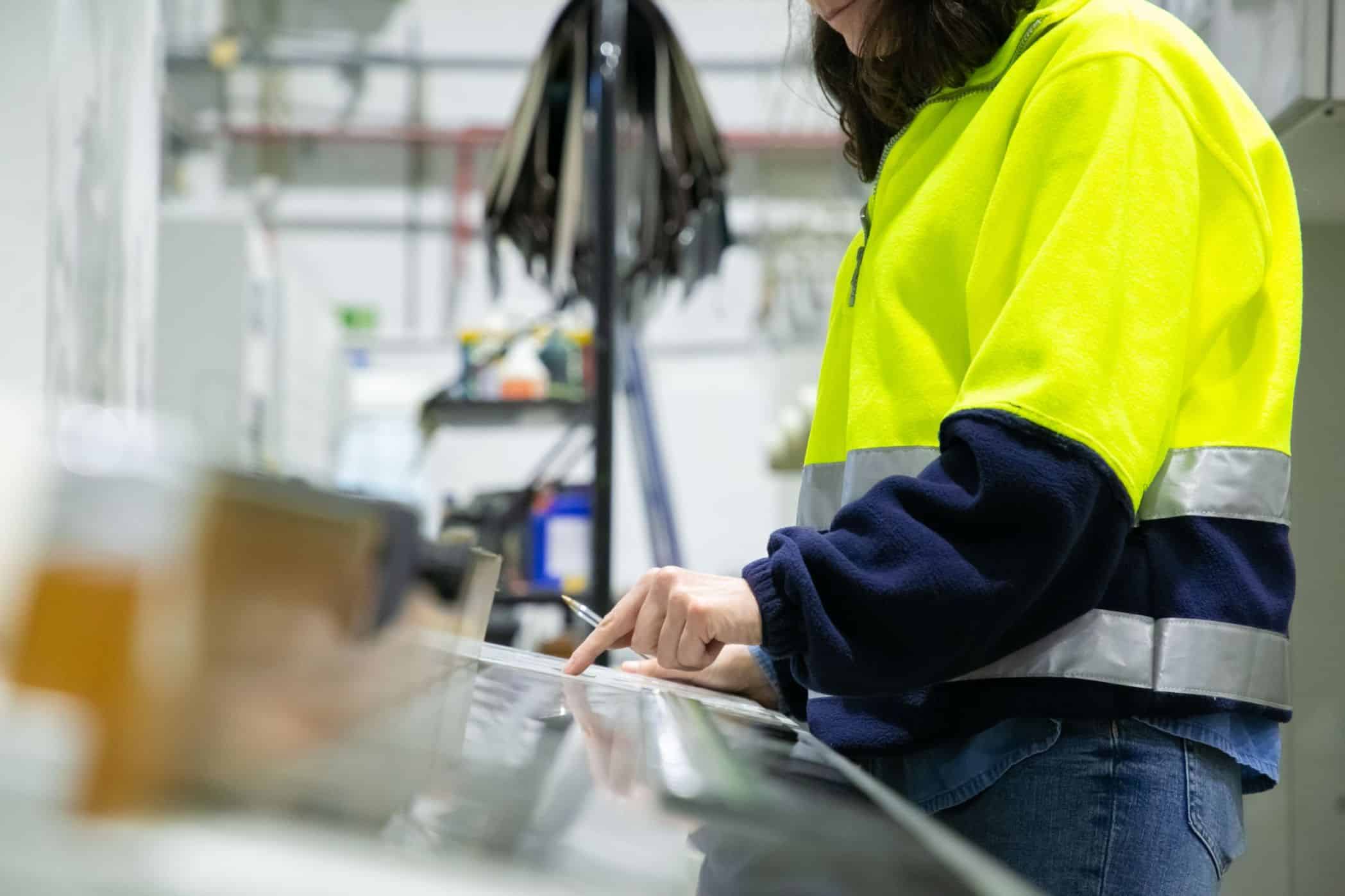 Trabalhadora em uma fábrica, vestindo uniforme de alta visibilidade, utilizando uma tela para acompanhar dados de produção.