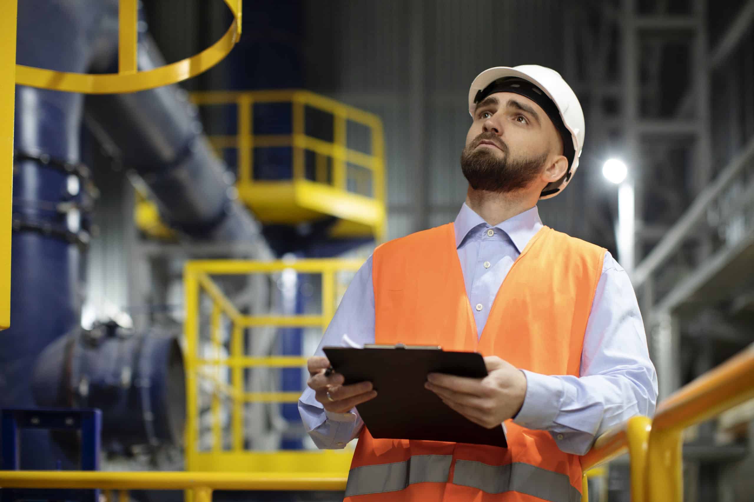 Engenheiro usando capacete branco e colete de segurança laranja, segurando uma prancheta enquanto inspeciona um ambiente industrial moderno com estruturas metálicas e tubulações ao fundo.