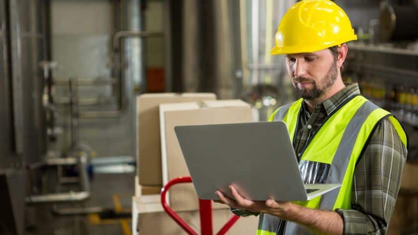 Homem vestindo capacete amarelo de segurança e colete refletivo segurando um laptop em um ambiente industrial, com caixas de papelão e tubulações ao fundo.