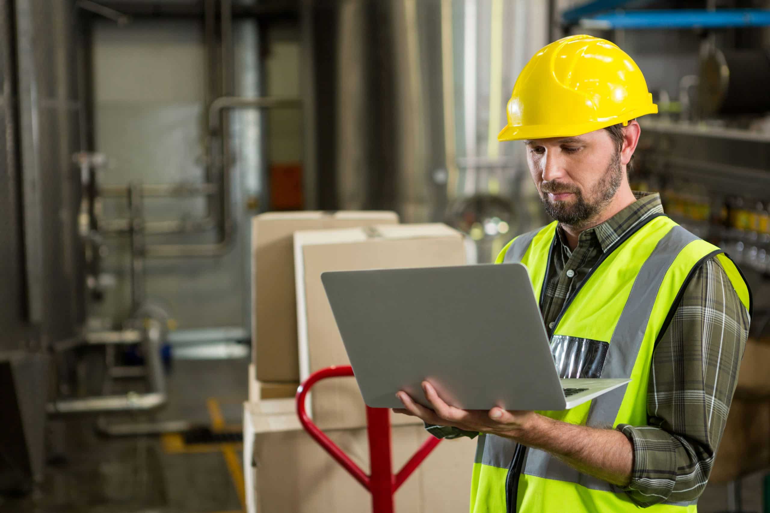 Homem vestindo capacete amarelo de segurança e colete refletivo segurando um laptop em um ambiente industrial, com caixas de papelão e tubulações ao fundo.