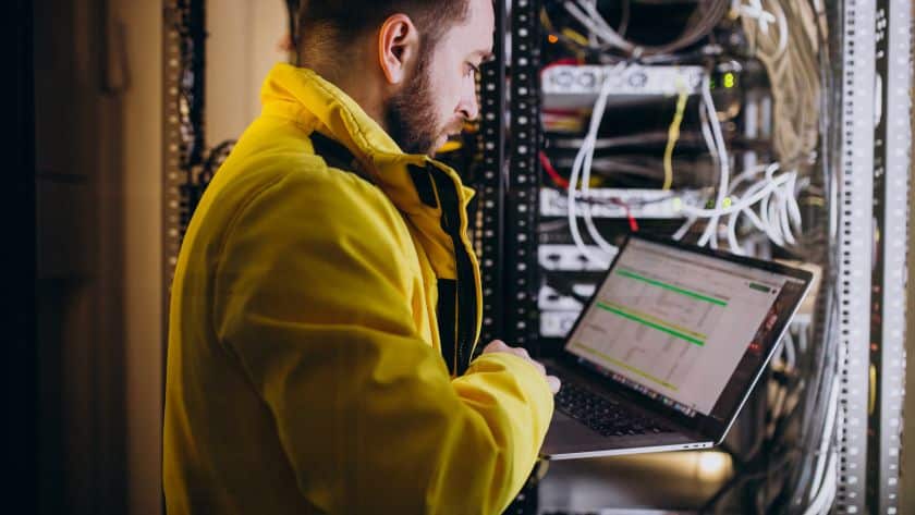 Homem jovem vestindo jaqueta amarela analisando dados em um laptop em frente a um rack de servidores com cabos conectados, em um ambiente tecnológico.