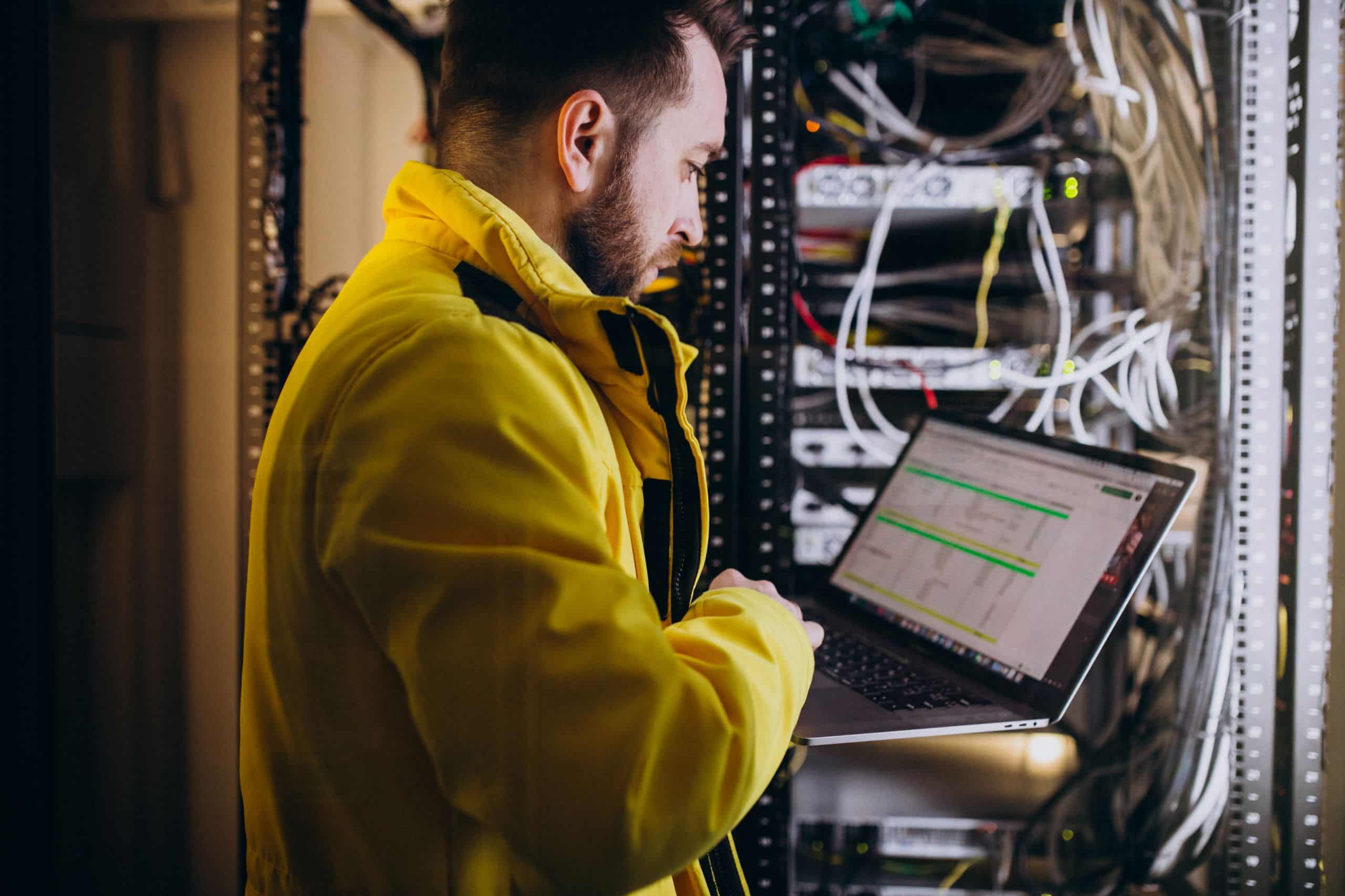 Homem jovem vestindo jaqueta amarela analisando dados em um laptop em frente a um rack de servidores com cabos conectados, em um ambiente tecnológico.