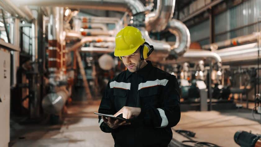 Trabalhador em uma planta industrial utilizando capacete amarelo, protetores auriculares e uniforme de segurança. Ele está operando um tablet em meio a tubulações e equipamentos industriais, indicando monitoramento ou execução de tarefas técnicas.