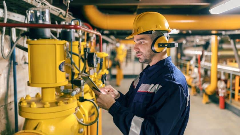 Trabalhador em uma usina utilizando capacete, protetores auriculares e uniforme de segurança. Ele está inspecionando máquinas industriais amarelas enquanto consulta um tablet, indicando monitoramento e controle de processos em um ambiente técnico