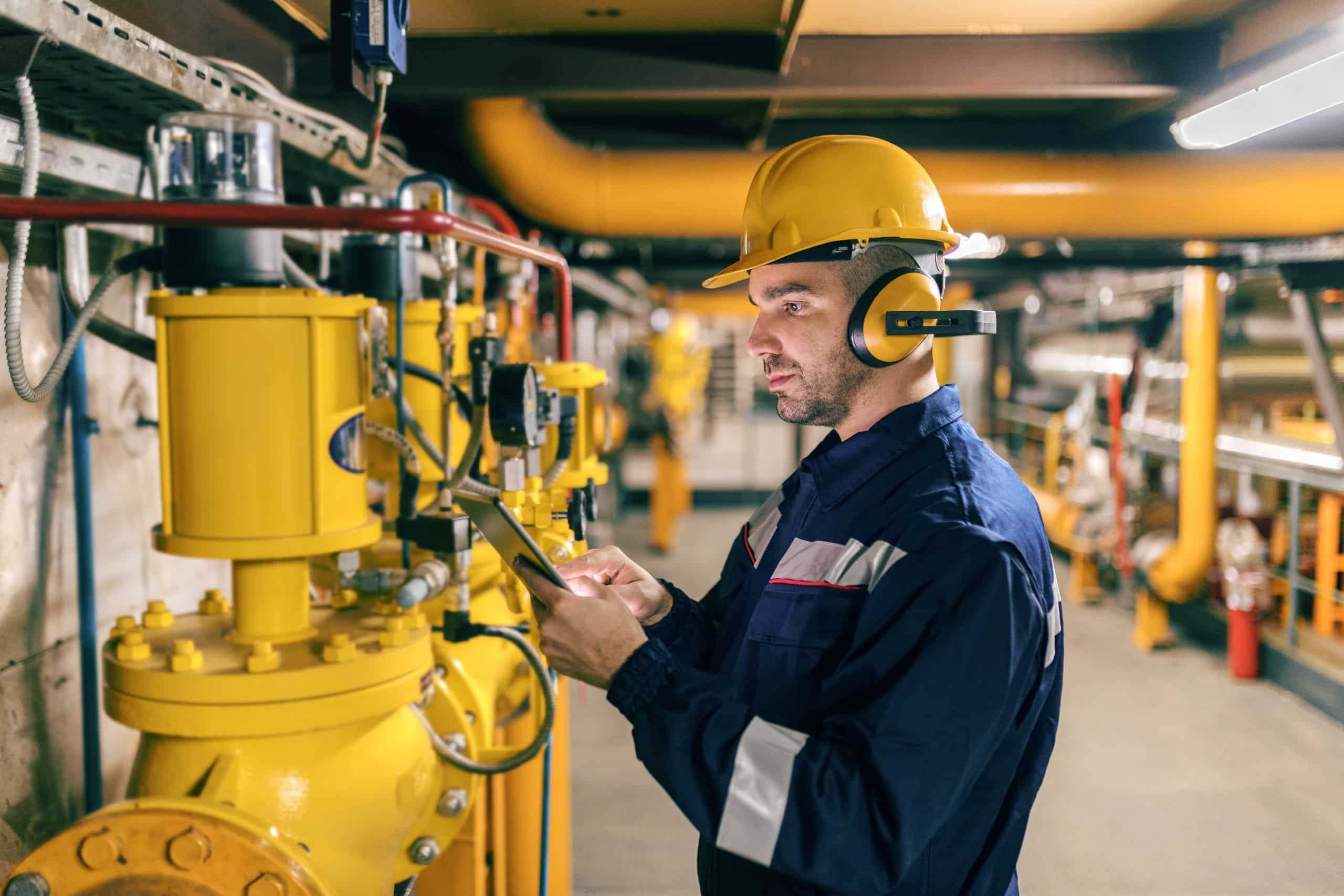 Trabalhador em uma usina utilizando capacete, protetores auriculares e uniforme de segurança. Ele está inspecionando máquinas industriais amarelas enquanto consulta um tablet, indicando monitoramento e controle de processos em um ambiente técnico