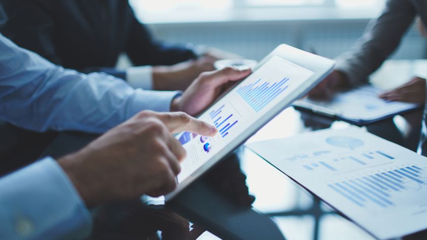 Pessoas em um ambiente corporativo analisando gráficos e dados financeiros em um tablet e em documentos impressos sobre a mesa. A cena sugere uma reunião de negócios ou uma discussão sobre gestão industrial e estratégia de mercado.