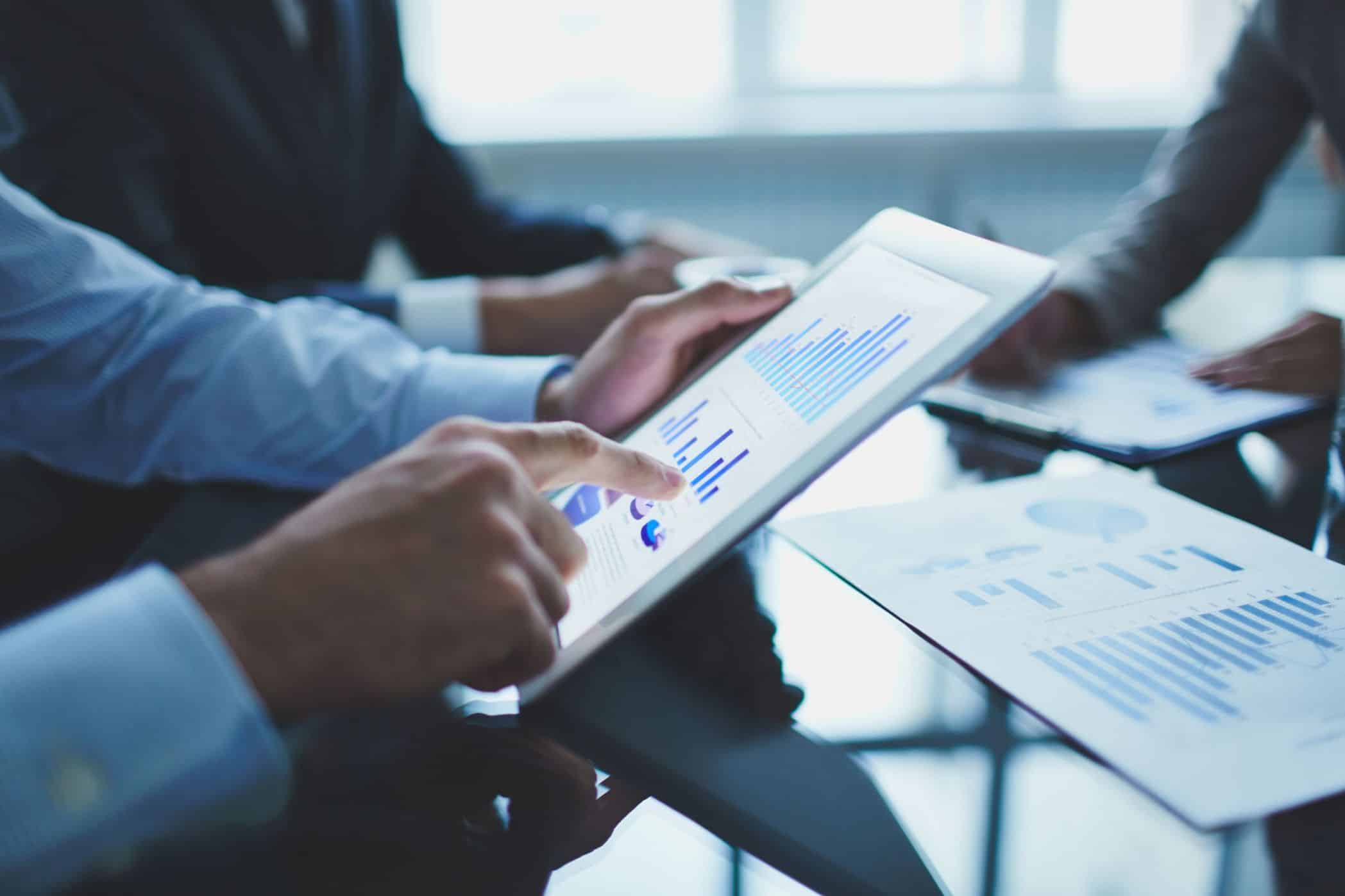 Pessoas em um ambiente corporativo analisando gráficos e dados financeiros em um tablet e em documentos impressos sobre a mesa. A cena sugere uma reunião de negócios ou uma discussão sobre gestão industrial e estratégia de mercado.