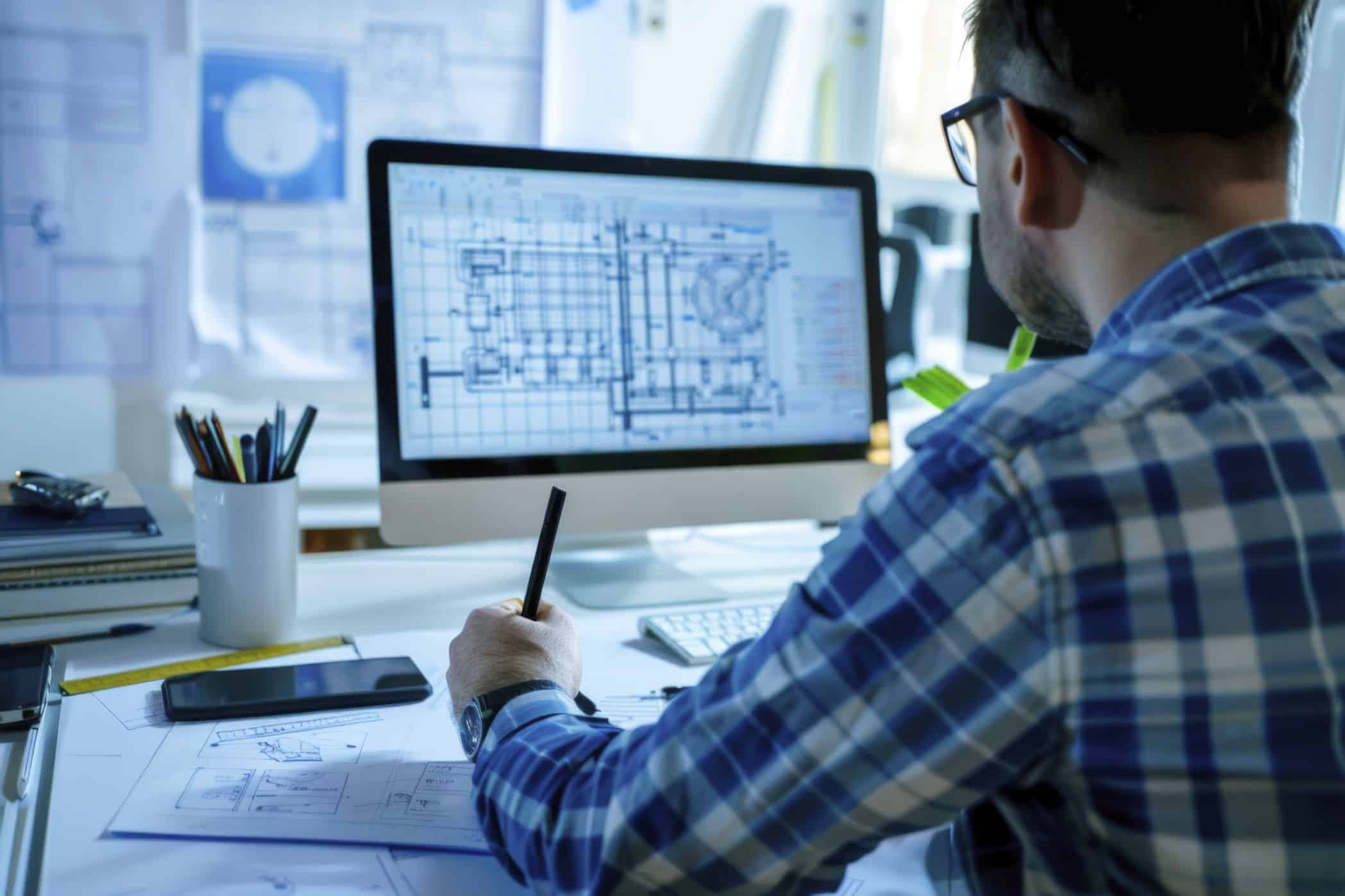 Engenheiro analisando um modelo digital de construção em um computador. O ambiente de trabalho está organizado com plantas arquitetônicas, régua e materiais de escritório. A tela exibe um projeto técnico detalhado.