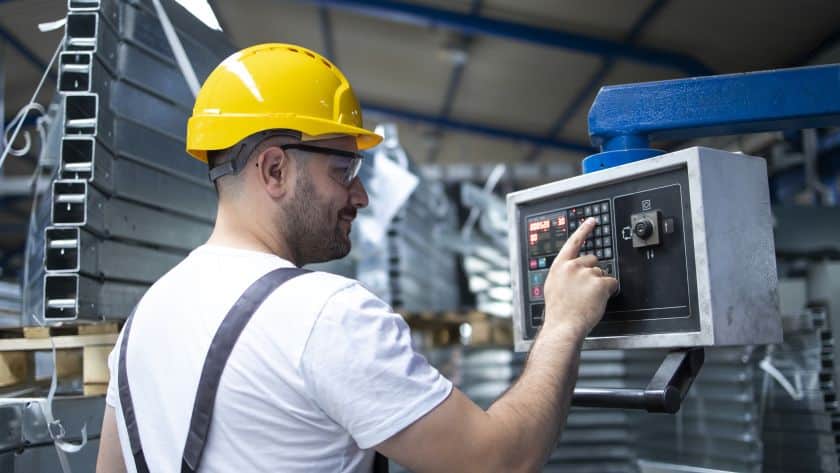 Trabalhador da indústria, vestindo capacete amarelo e óculos de proteção, opera um painel de controle eletrônico em um ambiente fabril. Ao fundo, estruturas metálicas e equipamentos industriais compõem o cenário.