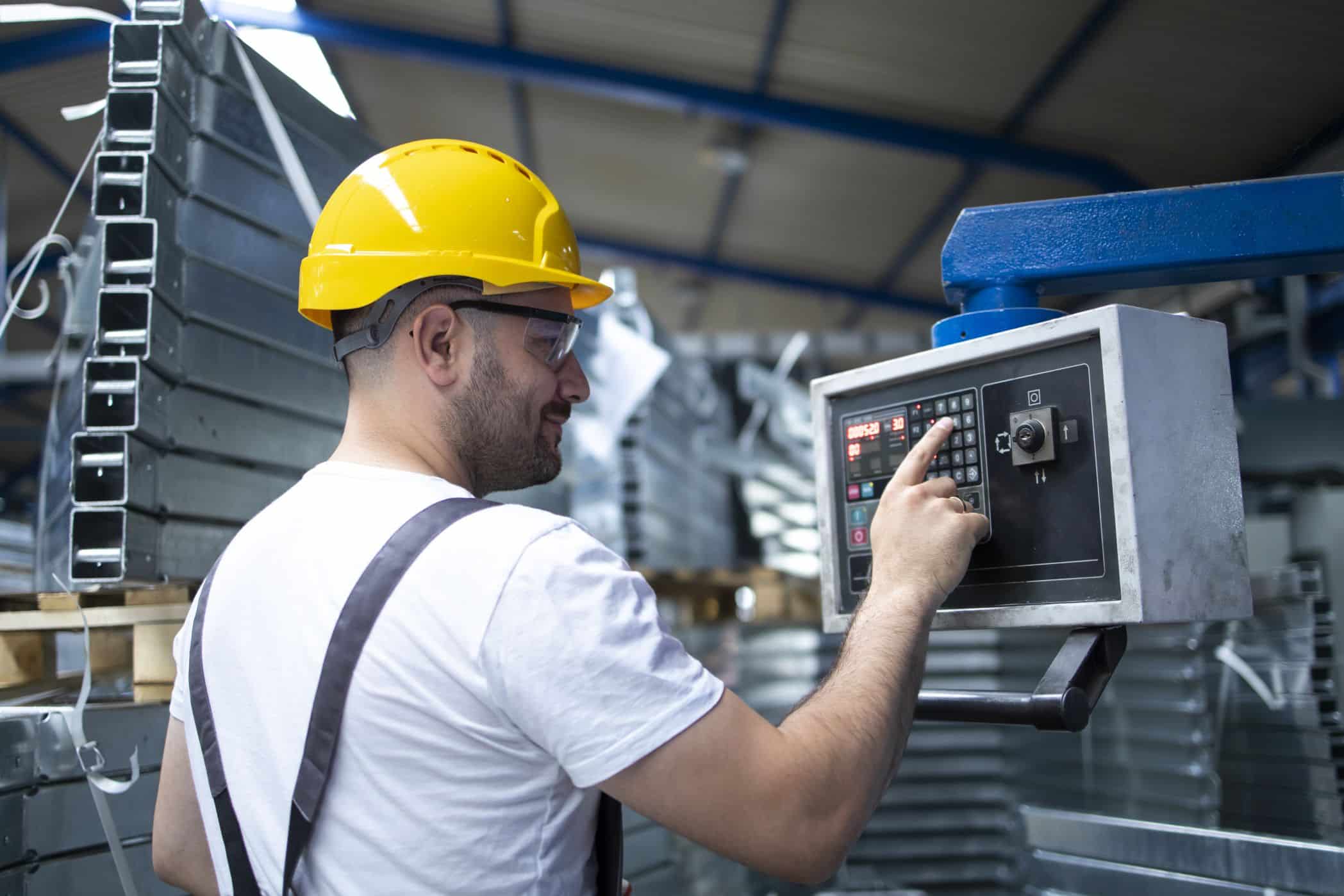 Trabalhador da indústria, vestindo capacete amarelo e óculos de proteção, opera um painel de controle eletrônico em um ambiente fabril. Ao fundo, estruturas metálicas e equipamentos industriais compõem o cenário.