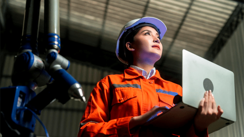 A imagem mostra uma mulher em um ambiente industrial, vestindo um macacão de segurança laranja com faixas refletivas e capacete branco, segurando um notebook enquanto observa algo à sua frente com expressão confiante. À esquerda da imagem, há parte de um braço robótico azul e cinza, sugerindo que ela está envolvida em atividades relacionadas à automação, engenharia ou manutenção tecnológica. O cenário ao fundo, com estrutura metálica e iluminação industrial, reforça o ambiente de fábrica ou centro de produção moderno, destacando o papel feminino na tecnologia e na indústria 4.0.