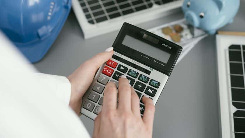 A imagem mostra uma pessoa usando uma calculadora grande, sugerindo que está realizando cálculos financeiros ou operacionais. Ao fundo, há elementos que remetem à temática de engenharia ou energia: dois painéis solares, um capacete de segurança azul e um cofrinho azul em formato de porquinho, do qual saem notas e moedas.