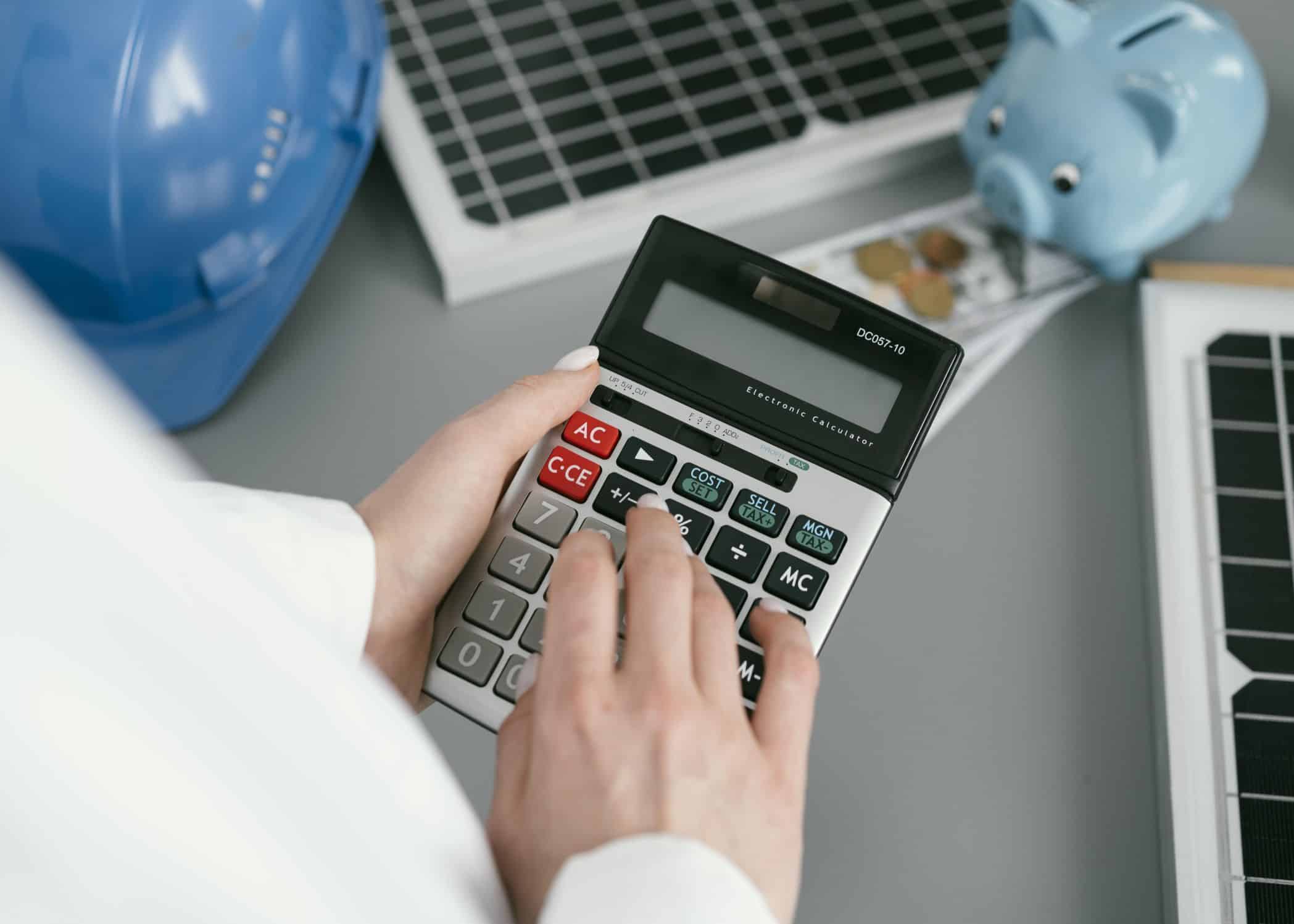 A imagem mostra uma pessoa usando uma calculadora grande, sugerindo que está realizando cálculos financeiros ou operacionais. Ao fundo, há elementos que remetem à temática de engenharia ou energia: dois painéis solares, um capacete de segurança azul e um cofrinho azul em formato de porquinho, do qual saem notas e moedas.