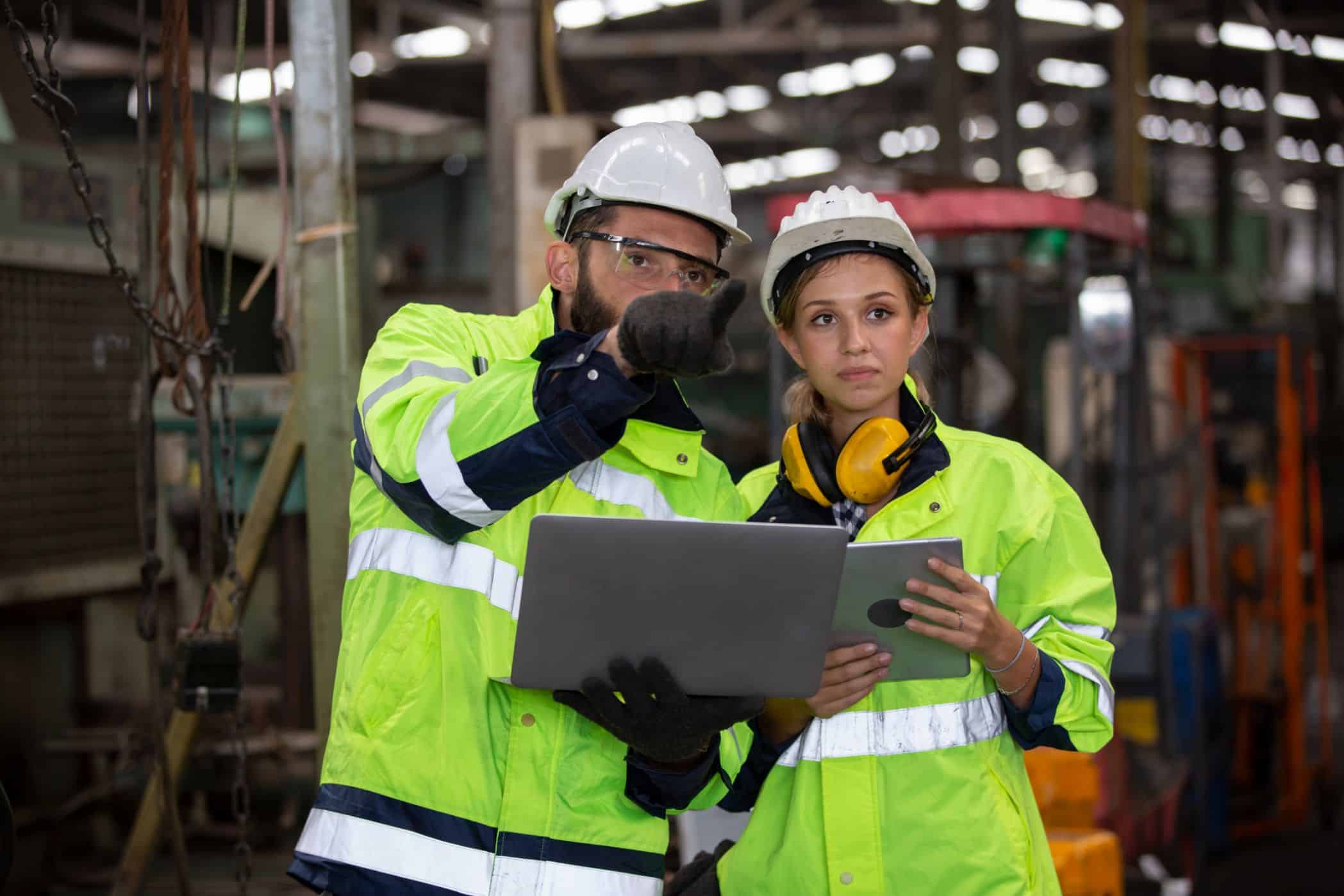 O ChatGPT disse: Dois trabalhadores usando equipamentos de segurança analisam dados em um laptop e tablet dentro de uma fábrica industrial.