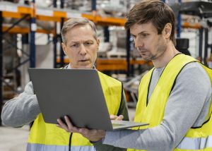 Dois trabalhadores com coletes refletivos analisando informações em laptop dentro de armazém.