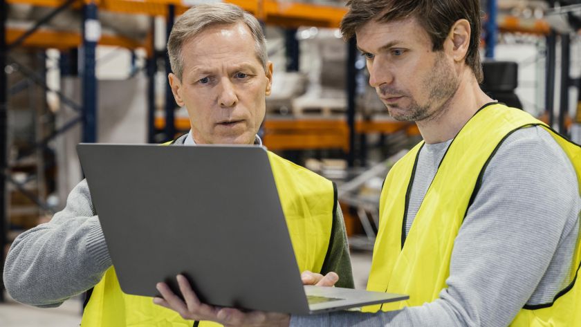 Dois trabalhadores com coletes refletivos analisando informações em laptop dentro de armazém.