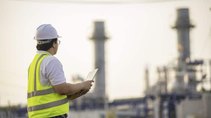 Engenheiro com capacete e colete refletivo usando tablet em área industrial.