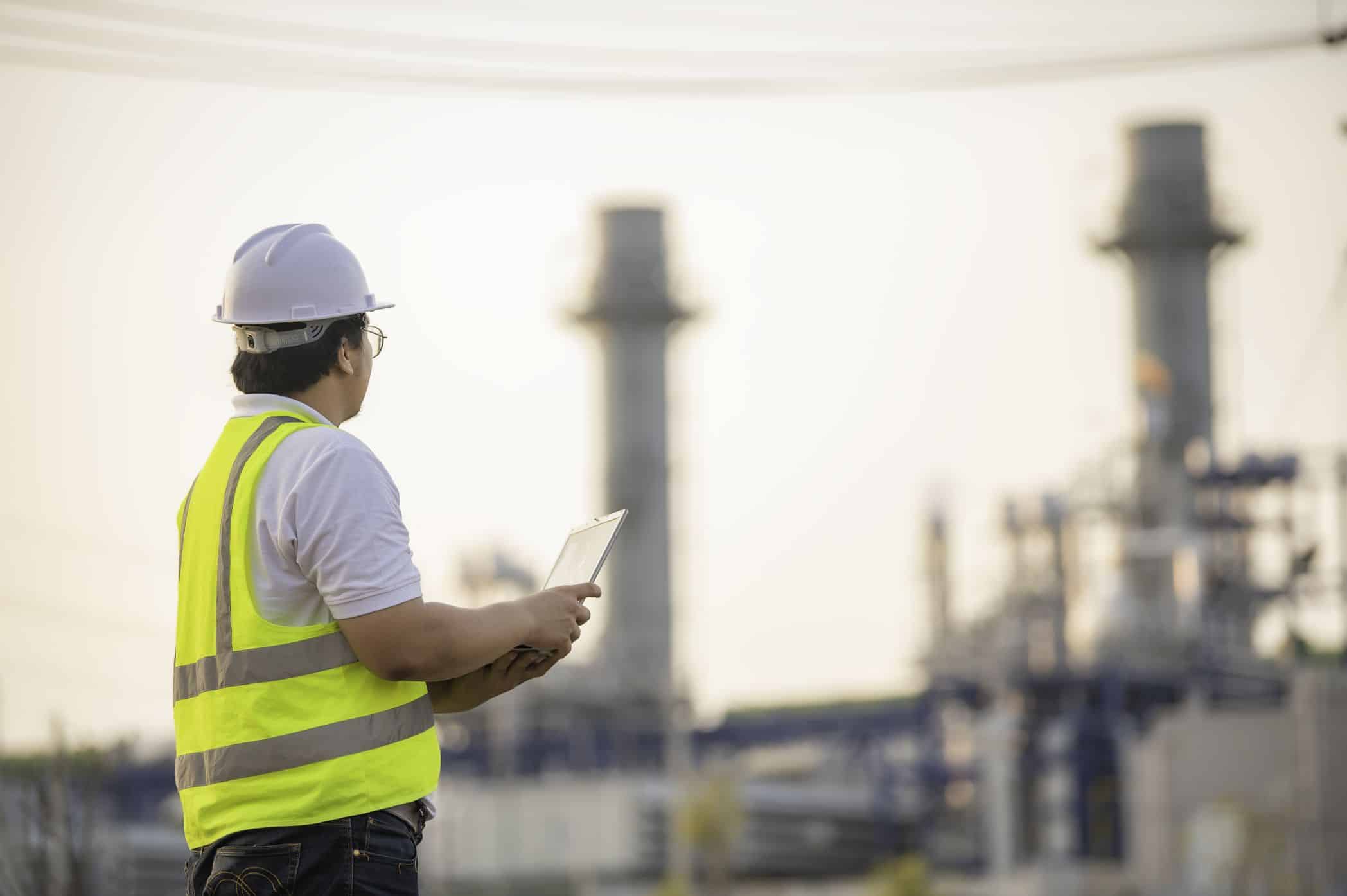 Engenheiro com capacete e colete refletivo usando tablet em área industrial.