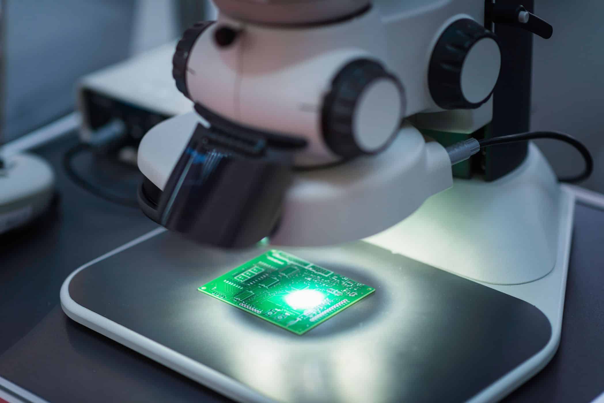 Microscópio analisando placa de circuito eletrônico iluminada.